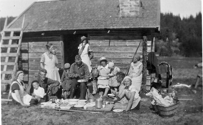 Bilden visar vuxna och barn som sitter utanför en liten stuga. På gräset ligger en filt där de har dukat upp mat