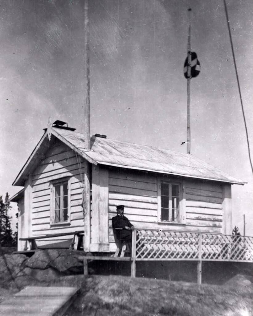 Bilden visar en äldre vitmålad stuga. Utanför stugan sitter en man med uniform