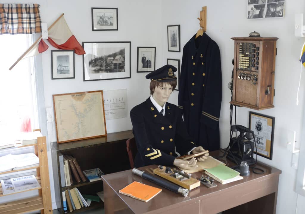 Bilden visar en skyltdocka klädd i lotsuniform som sitter vid ett skrivbord. Här syns också en gammal telefon.