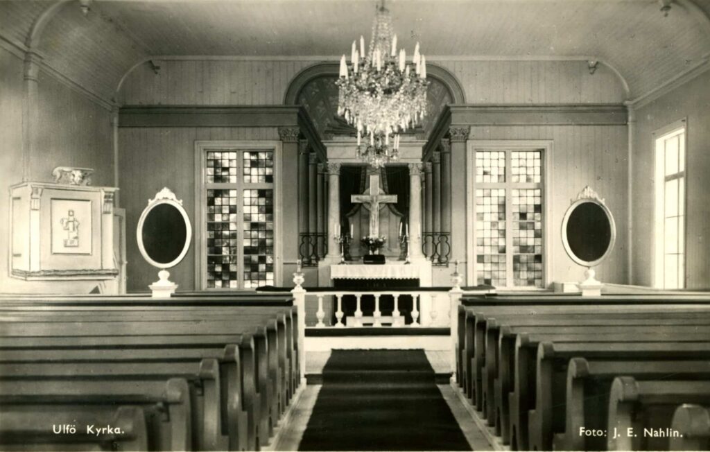 Bilden visar en interiör från Ulvö kyrka. Här finns kyrkbänkar, kristallkrona i taket, predikstol och altare
