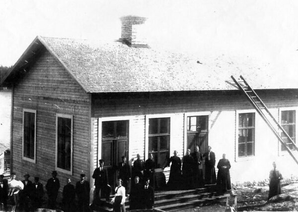 Bilden visar ett hus byggt av trä. Utanför huset står flera kvinnor och män