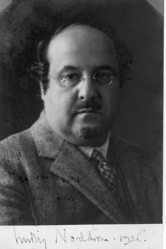 Bilden visar ett porträtt av en man med skägg, mustasch och glasögon